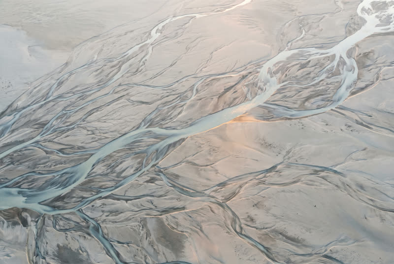 A braided river from above, many fine threads of water converging into one channel across pale sand