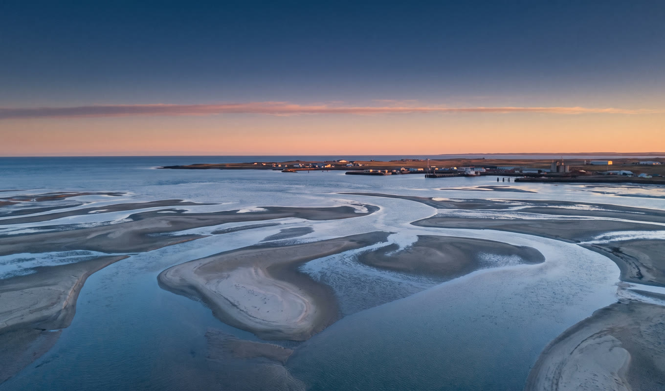 Low aerial over a tidal delta before sunrise, braided channels and sandbanks, a small working harbour catching the first light
