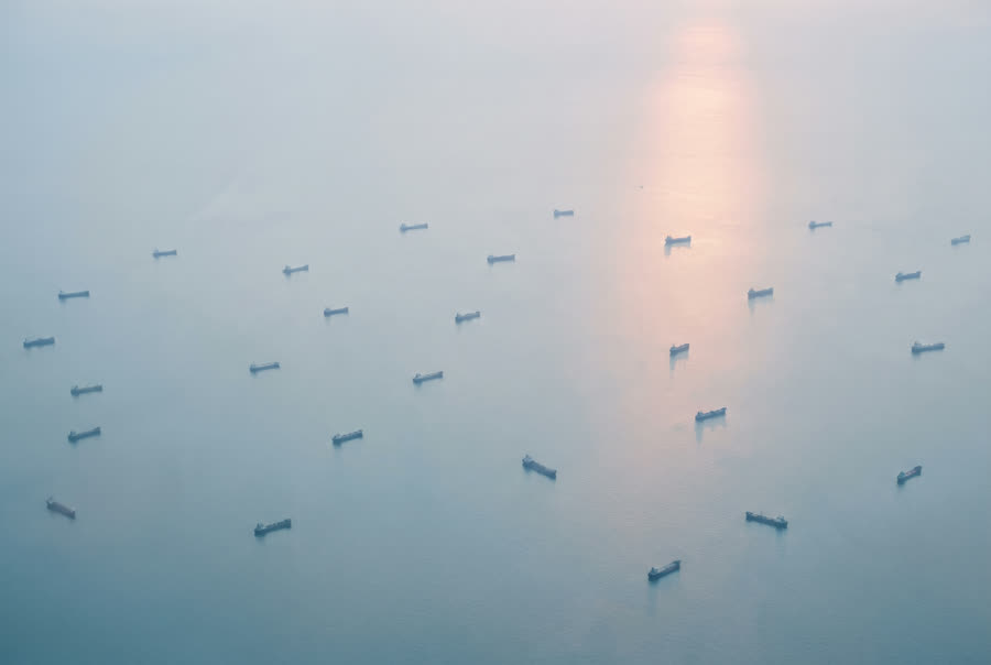 A wide anchorage at first light, many cargo ships lying at anchor on pale calm water