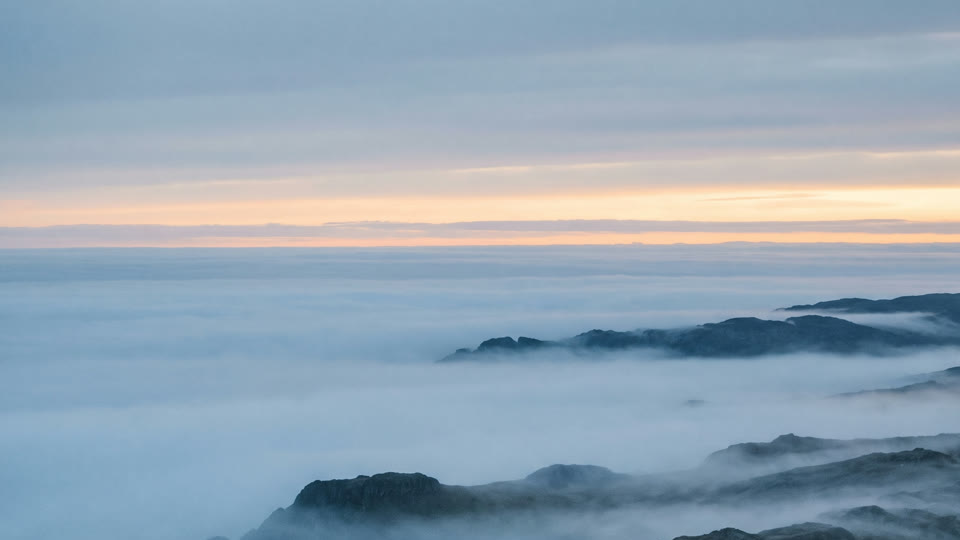 Low fog filling the valleys between dark hills, under a pale dawn sky.