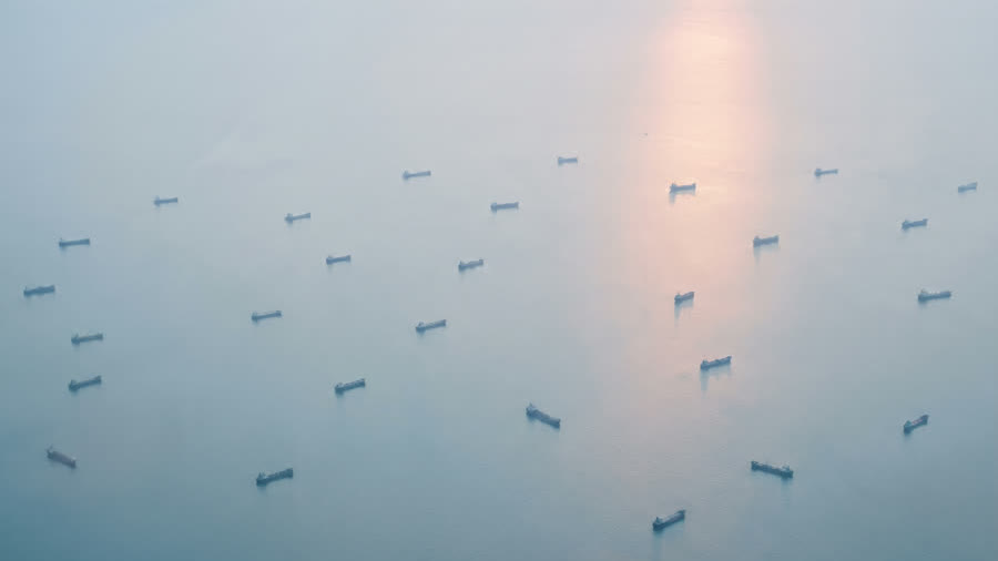 Ships at anchor on pale, calm water, with sunlight reflecting off the sea.