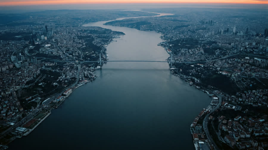 A bridge across a wide strait between two cities at dusk, seen from above.