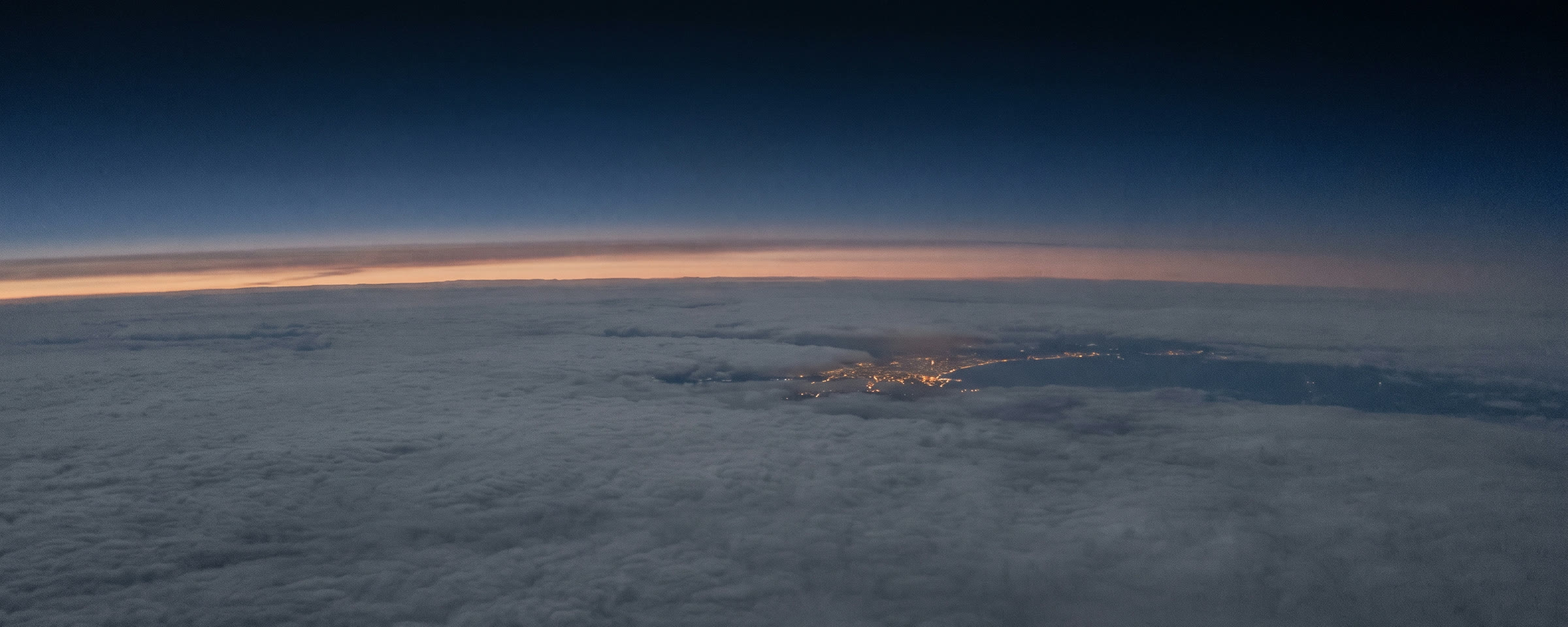 View from high altitude before sunrise, a flat cloud deck below and the lights of a coastal city showing through a break in the cloud