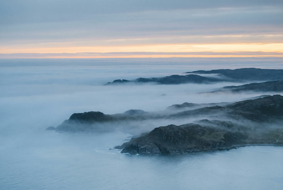 Sea fog at dawn erasing a low coastline, only the highest headlands showing through, one thin band of warm light at the horizon