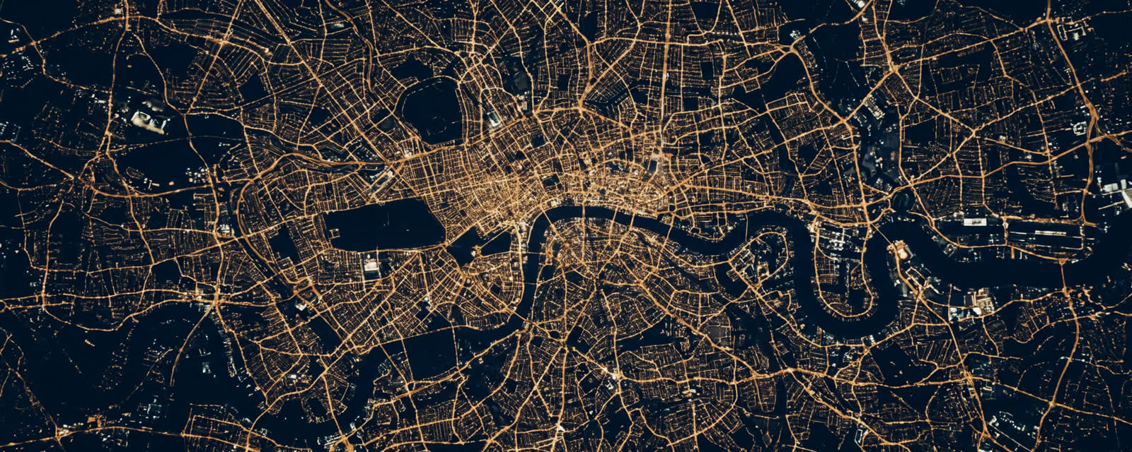 A city at night from very high altitude, streets reading as a web of warm light across dark ground, a river cutting through as a black void